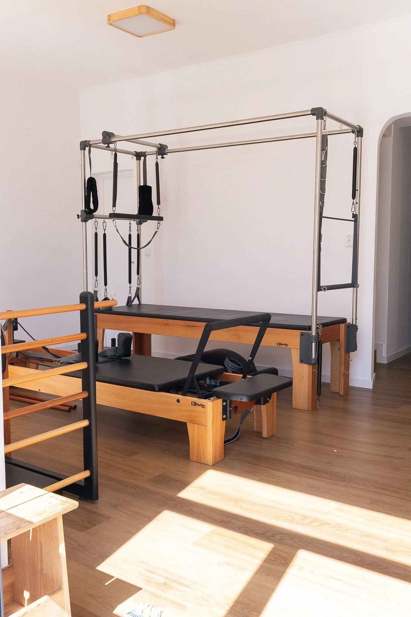 Sala de fisioterapia com equipamentos de suporte para exercícios, incluindo uma estrutura de metal com faixas de resistência e uma mesa de pilates de madeira com estofado preto.