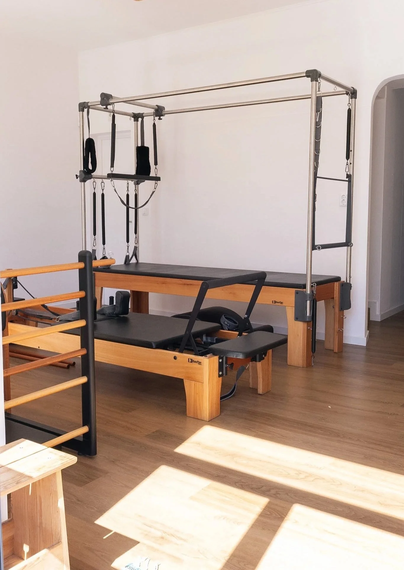 Sala de fisioterapia com diferentes equipamentos de exercícios, incluindo uma estrutura de barras de metal com várias faixas e argolas, além de um aparelho de pilates em um piso de madeira clara.