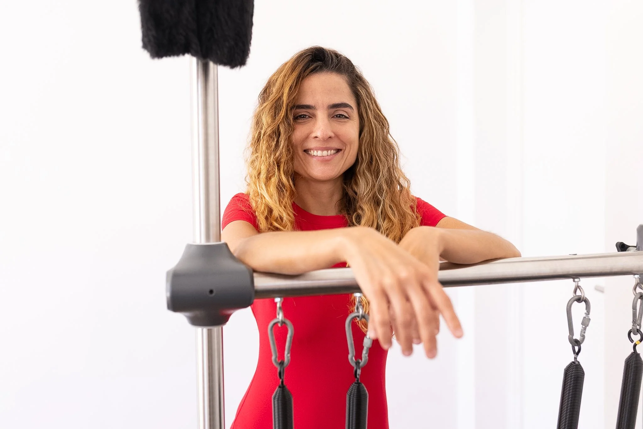 Mulher sorridente posicionada entre equipamentos de Pilates, usando uma blusa vermelha, com braços apoiados em uma estrutura de Pilates com molas.