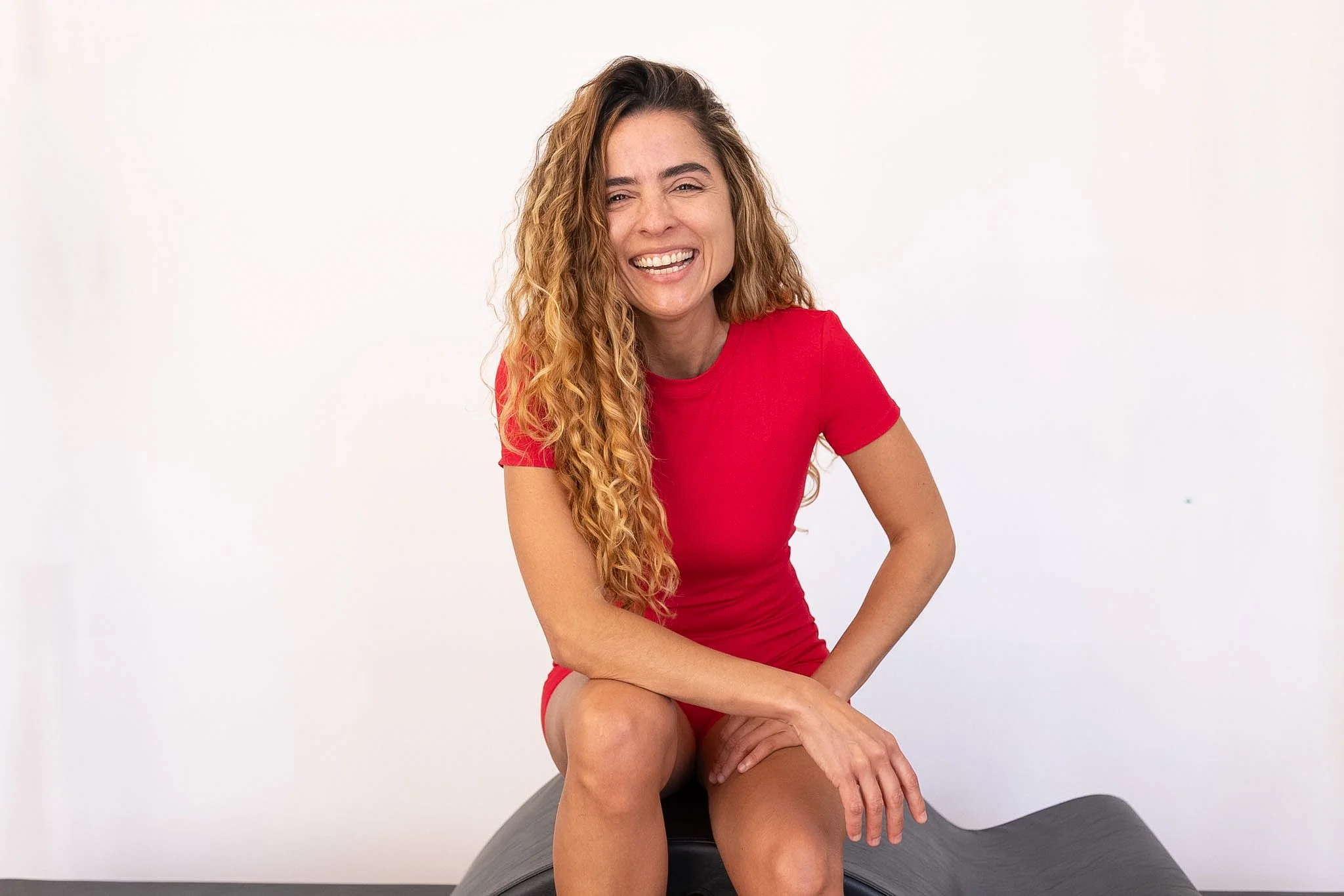 Mulher sorridente com cabelo encaracolado, vestindo camiseta vermelha, sentada sobre um objeto de cor escura, contra fundo branco.