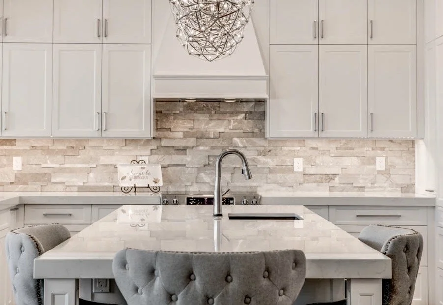 Modern kitchen with white cabinets, a white marble island, gray tufted chairs, a chrome faucet, and a stone backsplash, with a decorative hanging light fixture.
