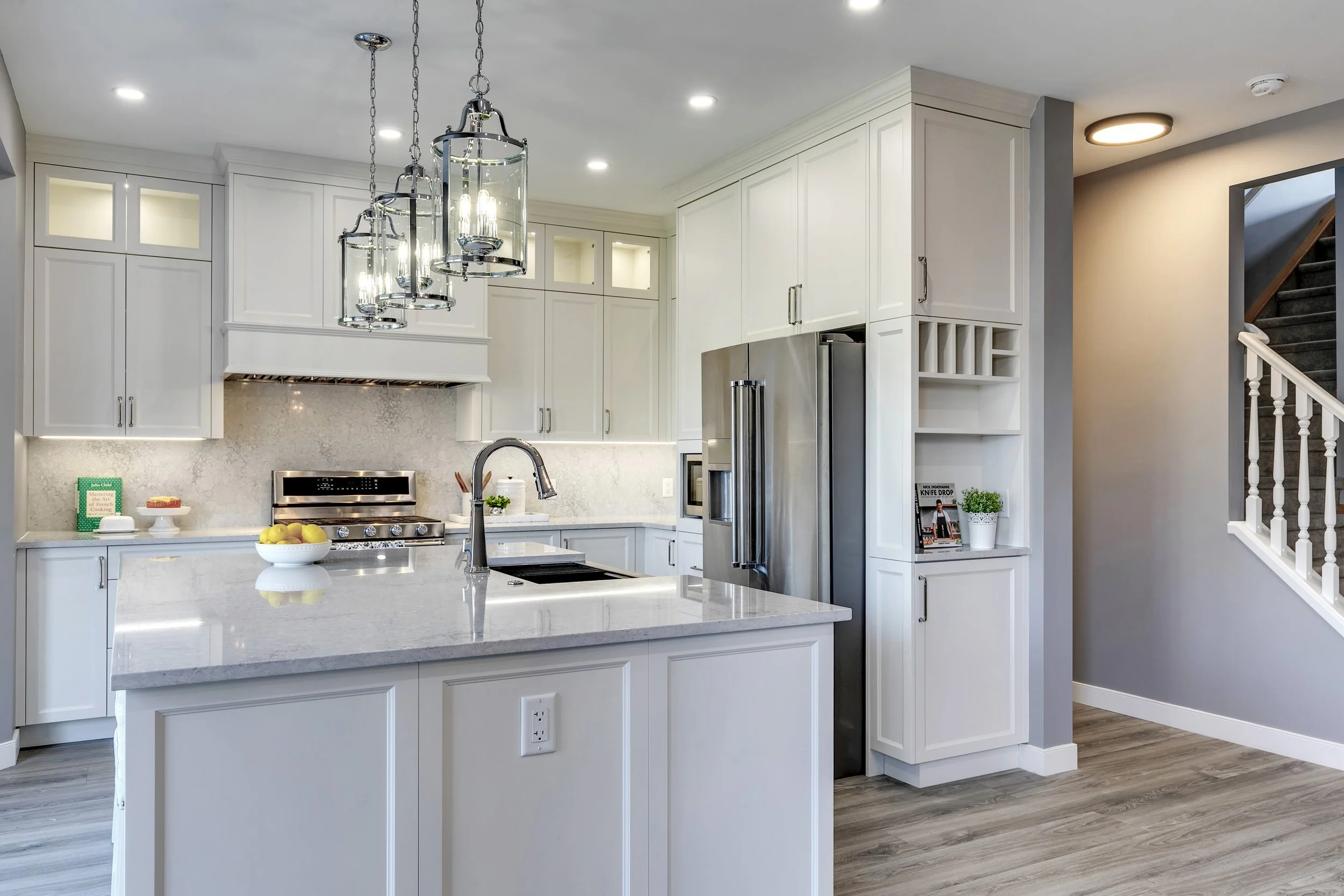 Modern white kitchen with island, pendant lights, stainless steel refrigerator, and staircase to the right.