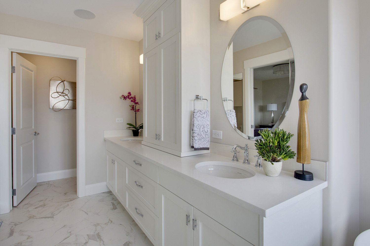 Modern bathroom with white vanity, large round mirror, small sink, potted plant, decorative mannequin, and towels.