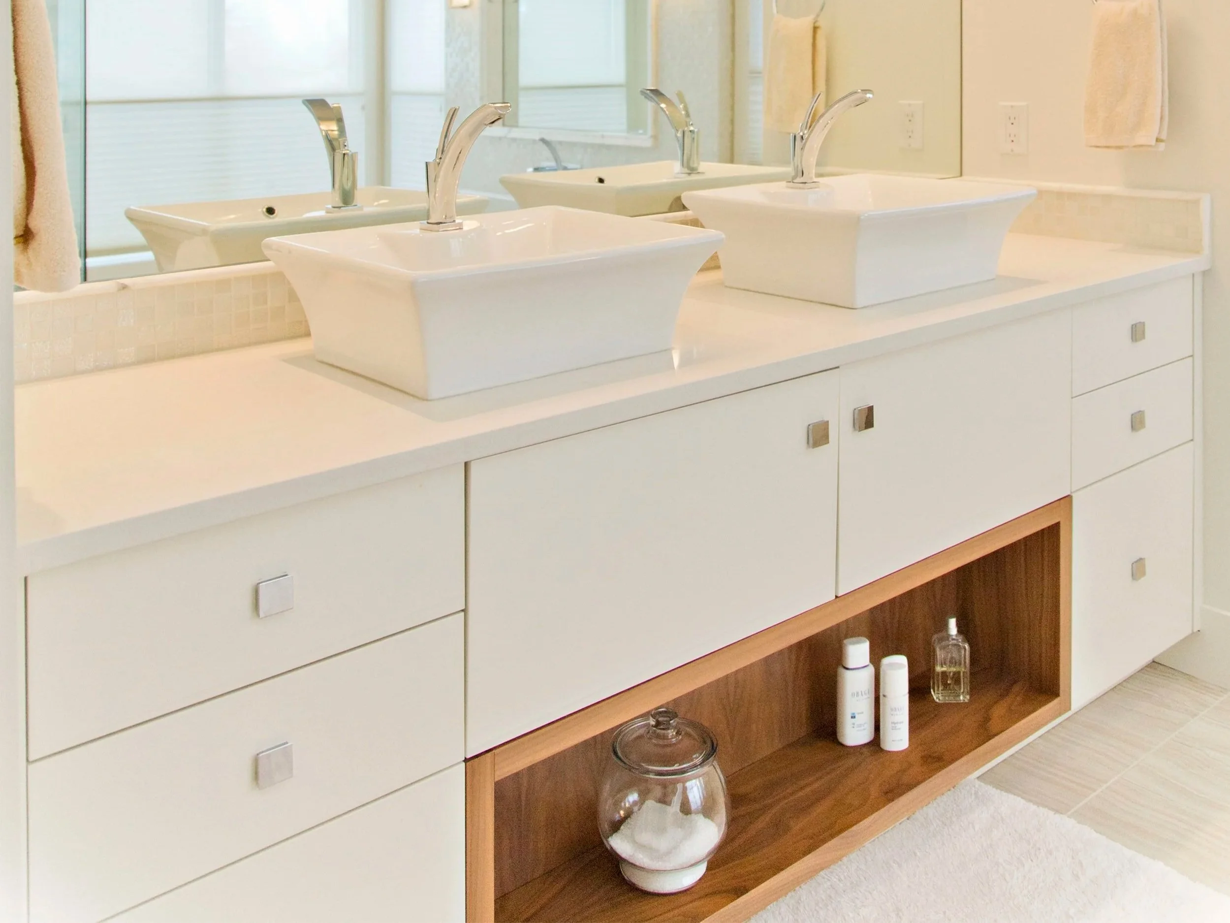 A bathroom vanity with three square vessel sinks and three chrome faucets, large mirror above, and a wooden shelf underneath holding bottles and a large glass jar.