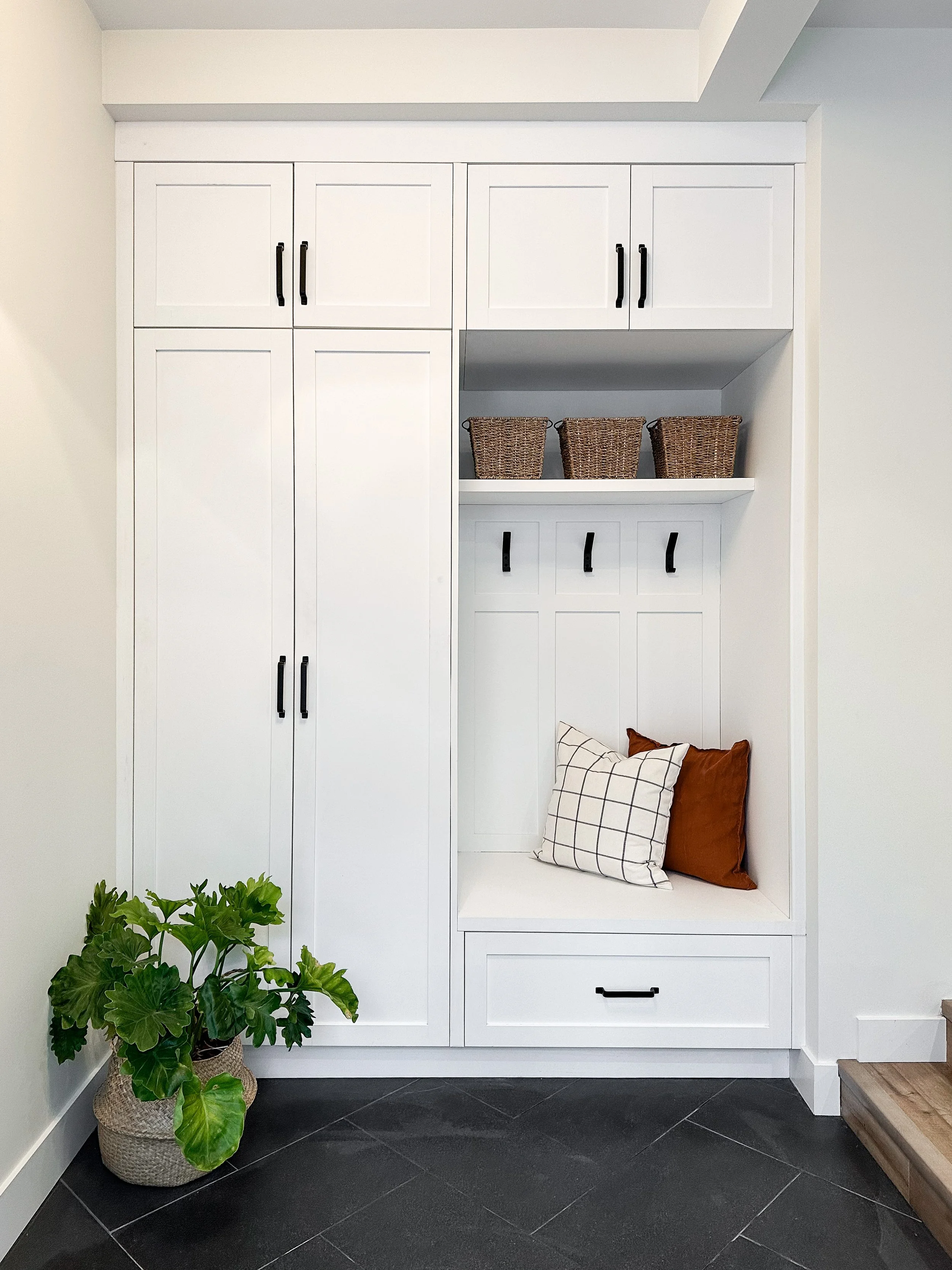 White built-in cabinet with black handles, a shelf with woven baskets, and a cozy nook with pillows, next to a potted green plant on a dark tiled floor.