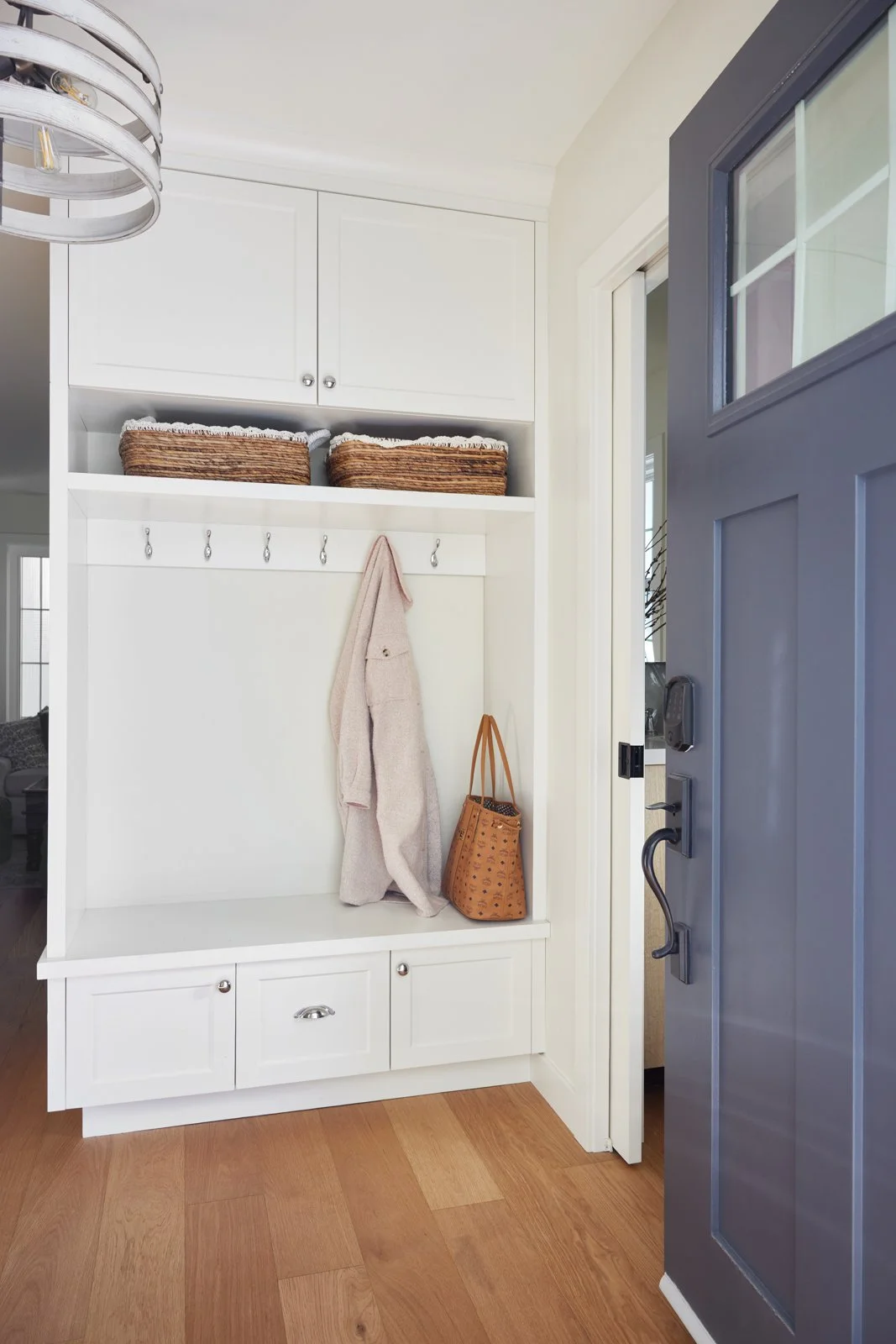 Entryway with a white mudroom bench featuring hooks with a beige coat hanging and a tan handbag on the floor. Overhead storage with woven baskets and white cabinets. Part of a gray front door with window panes is visible.