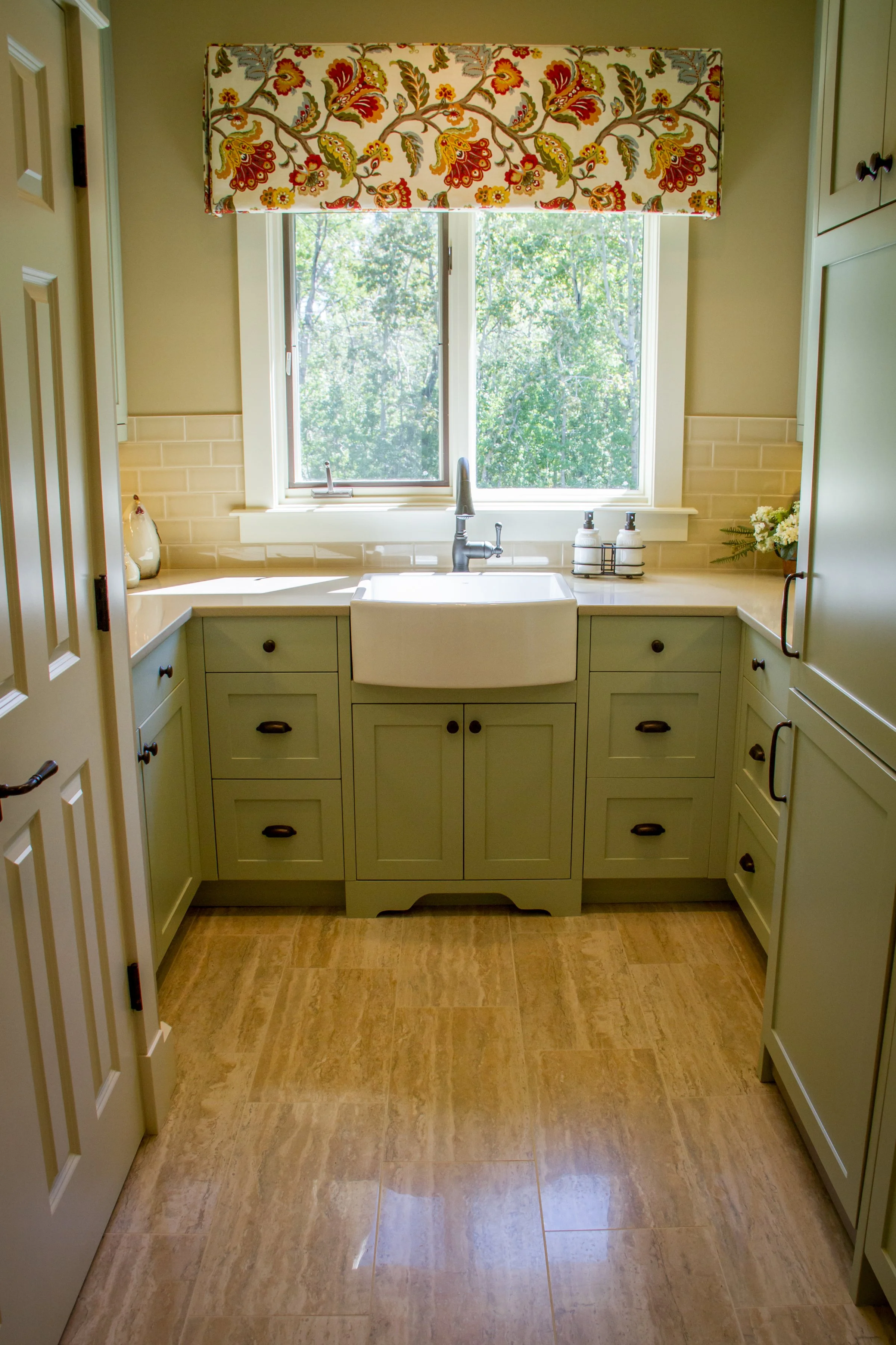 A cozy kitchen sink area with a farmhouse-style white apron sink, a modern faucet, and a large window. The window has a colorful floral valance and looks out onto a green, leafy view. The surrounding cabinetry is painted in a light green color, and t