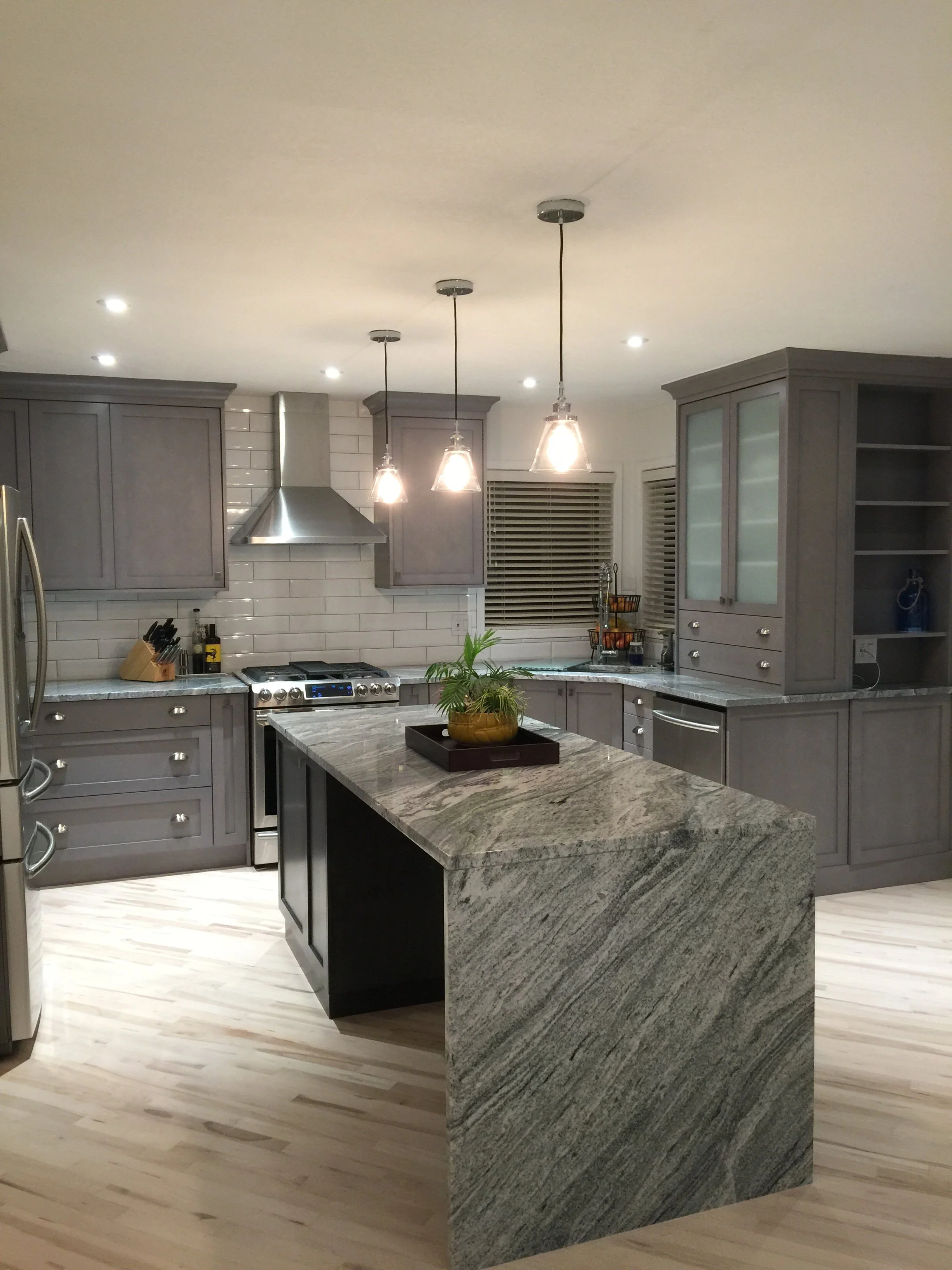 Modern kitchen with gray cabinets, a granite island, pendant lights, white subway tile backsplash, and stainless steel appliances.
