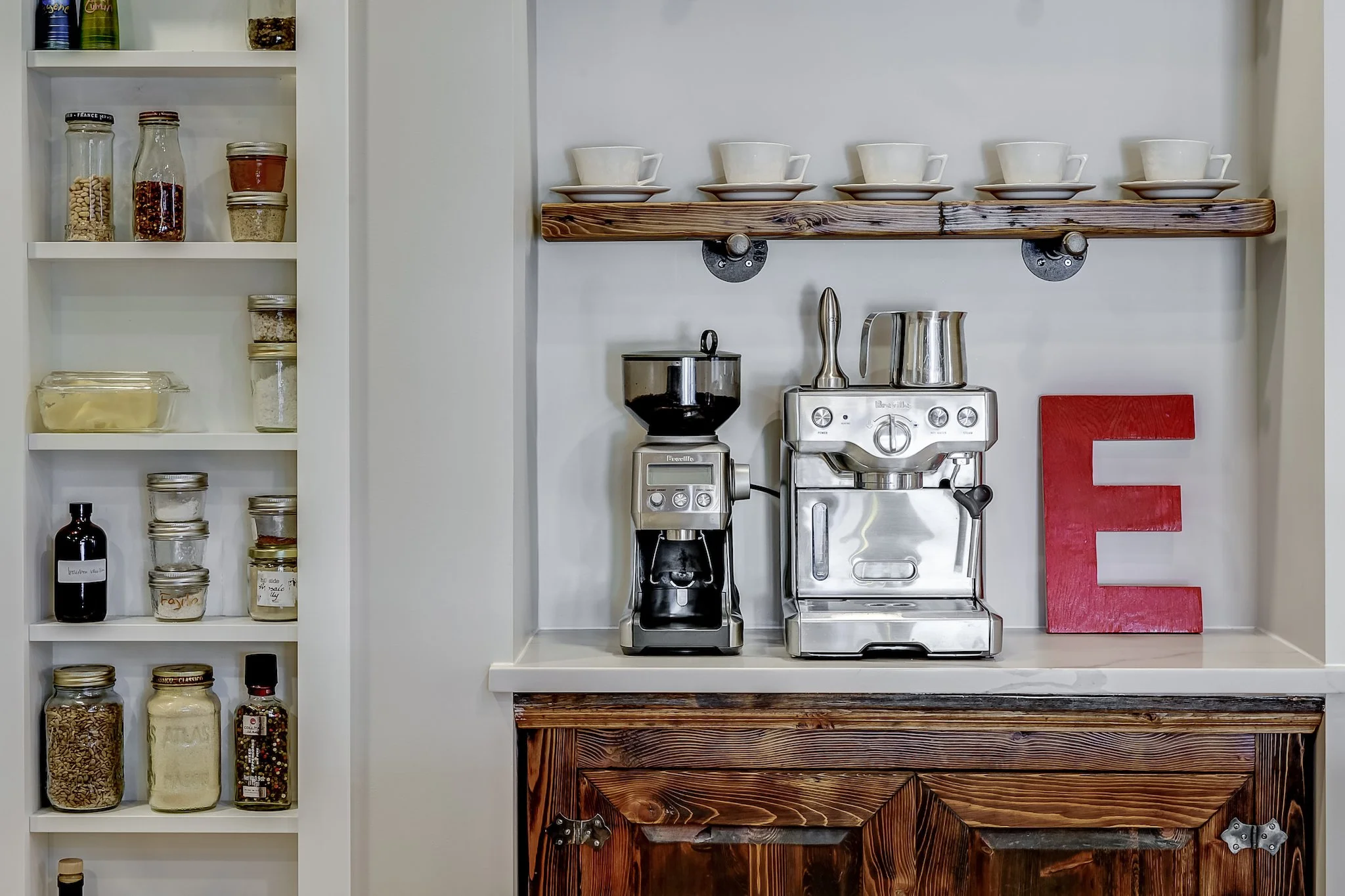 A kitchen corner featuring three white teacups on matching saucers on a wooden shelf, a coffee grinder, espresso machine, and the letter E in red on a white counter, with a white open shelving unit filled with jars of spices and ingredients.