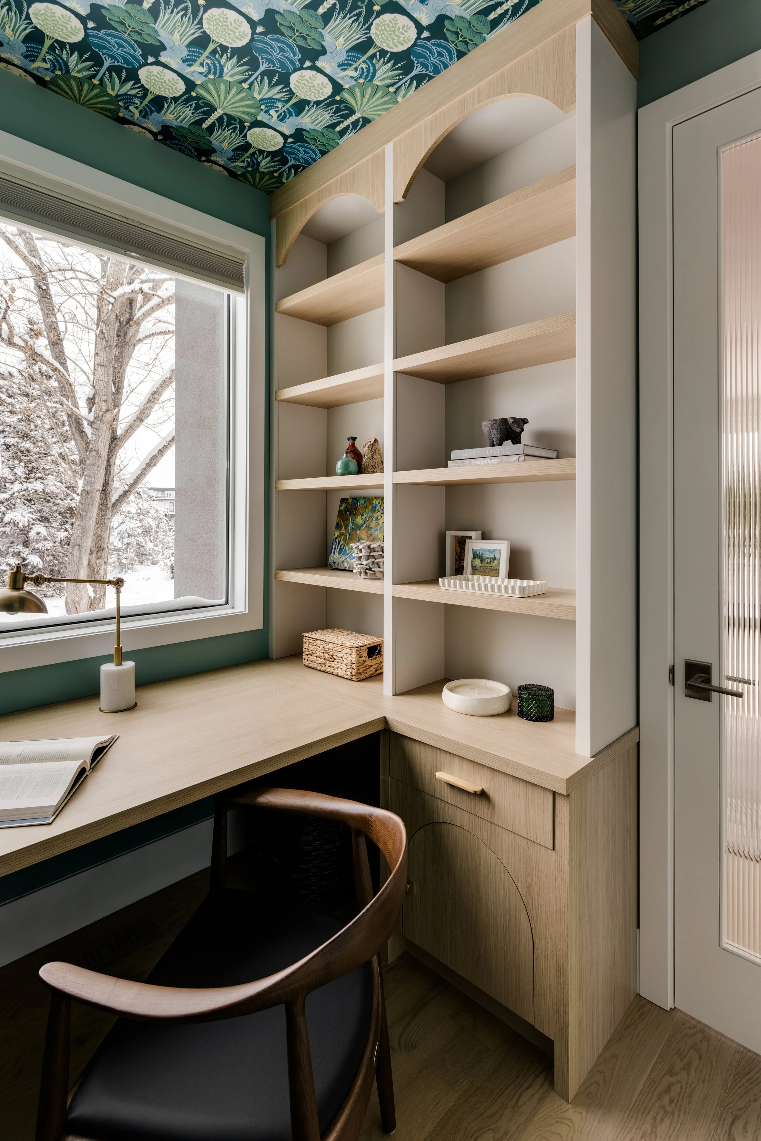 A cozy workspace with a wooden desk, black and wooden chairs, large window revealing a snowy landscape, and a built-in bookshelf with decorative items and books.