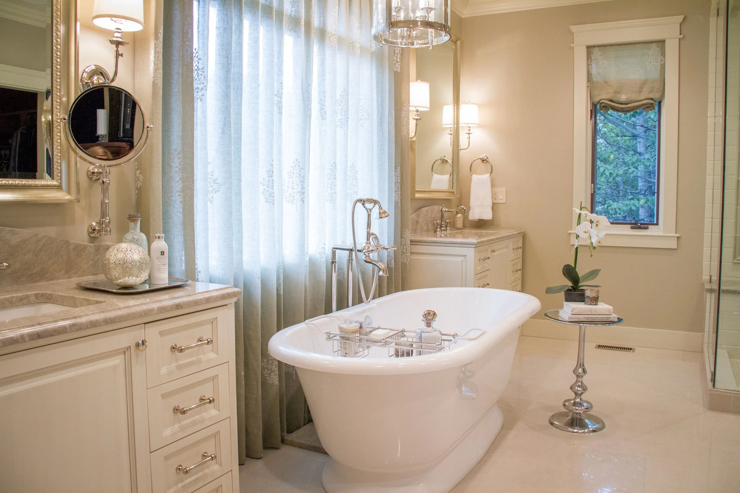 A luxurious bathroom with a freestanding bathtub, a large mirror, light-colored cabinetry, a small table with books and a flower, a window with sheer curtains, and multiple wall-mounted lamps.