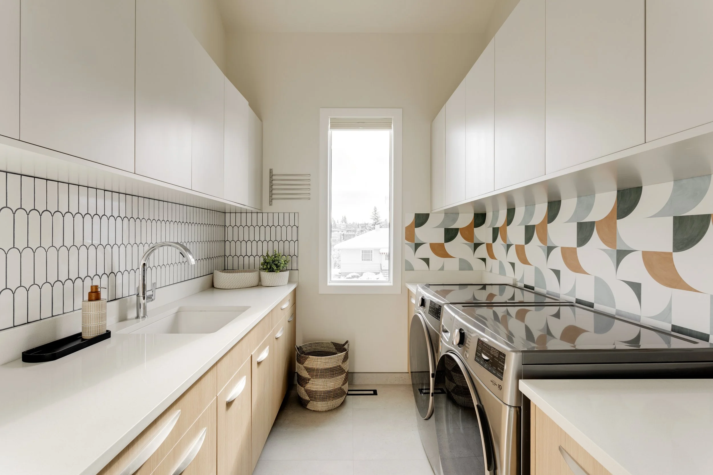 A modern laundry room with white cabinetry, patterned tiles behind the sink and washing machine, and a large window letting in natural light.