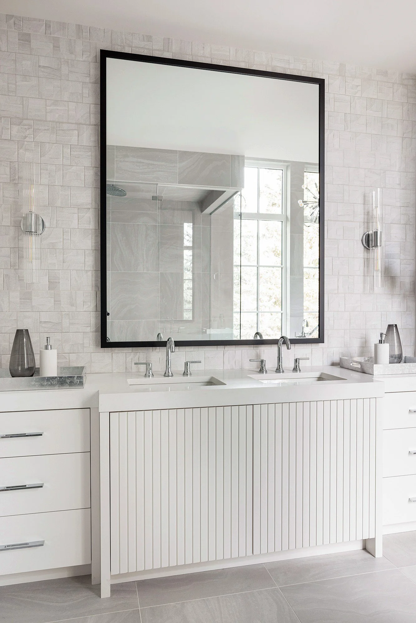 Modern bathroom with double sink vanity, large mirror, light gray textured wall tiles, and two standing lamps on marble trays.