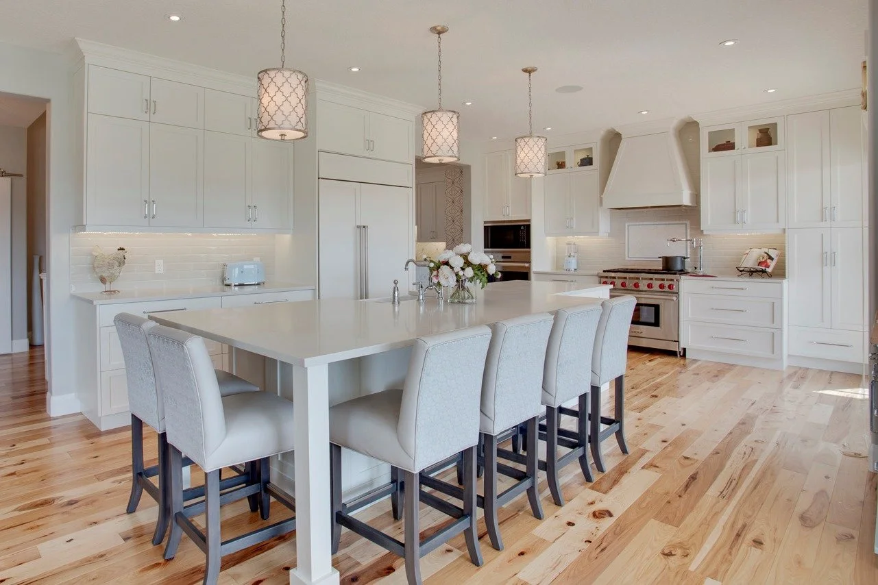 Bright white kitchen with a large island, beige barstools, wooden floors, pendant lights, and modern appliances.