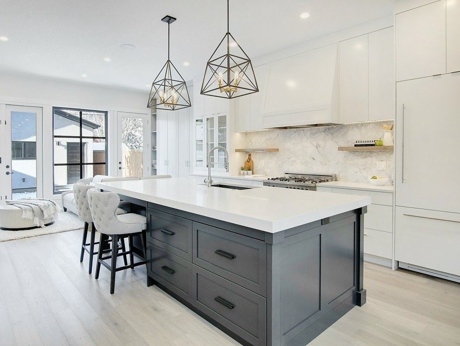 Modern kitchen with white cabinets, a marble backsplash, and a central island with dark gray drawers. Pendant lights hang above the island. There are barstools with white upholstery at the island. Sunlight streams through large glass doors leading to