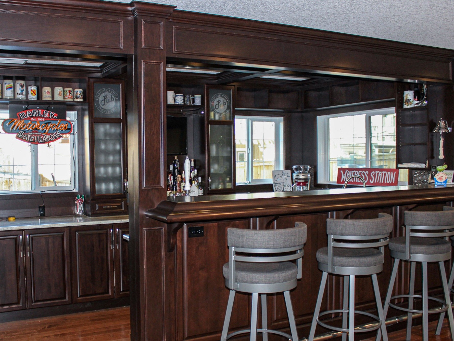 A home bar area with dark wood cabinetry, a marble countertop, three beige barstools, and windows behind. Signs read 'Harley's Motorcycles' and 'Waitress Station' are visible.