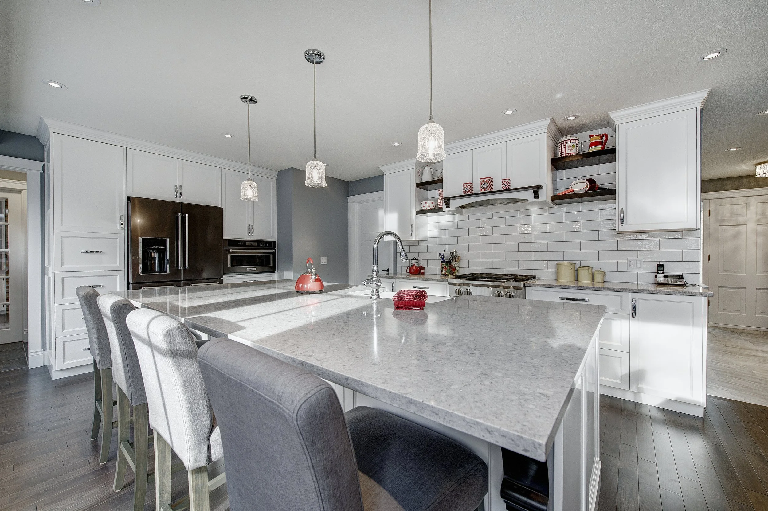 A modern kitchen with white cabinets, a large kitchen island with a granite countertop, pendant lights, and stainless steel appliances. There are chairs around the island, and decorative red and white kitchenware on open shelves.