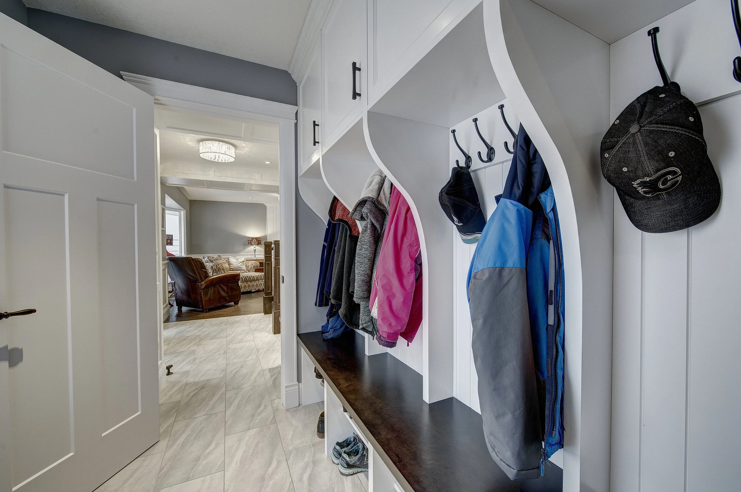 Entryway with white built-in hooks holding jackets, hats, and shoes, leading to a living room with a sofa and a chandelier.