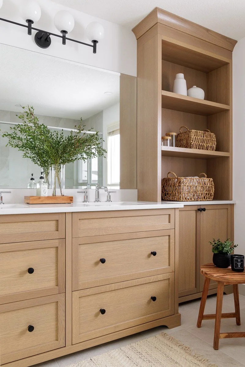 Bathroom vanity with a wooden cabinet, marble countertop, and a large mirror with a plant and soap dispensers in front. Open wooden shelving with baskets and decorative items. Small side table with a potted plant and a small device.