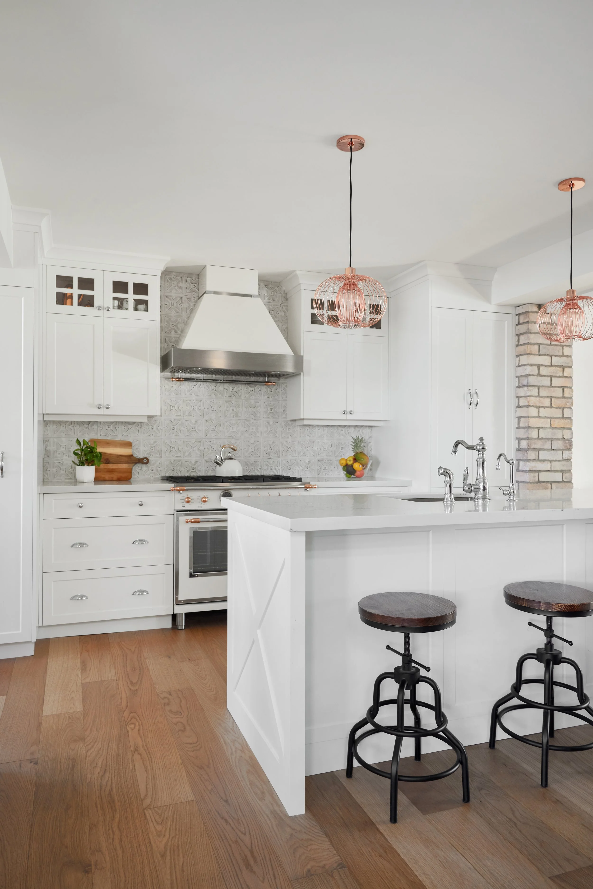 A modern kitchen with white cabinets, a stainless steel range hood, and a white island with two black and wood bar stools. The kitchen has a wooden floor, pendant lights with rose gold accents, and a brick accent wall.