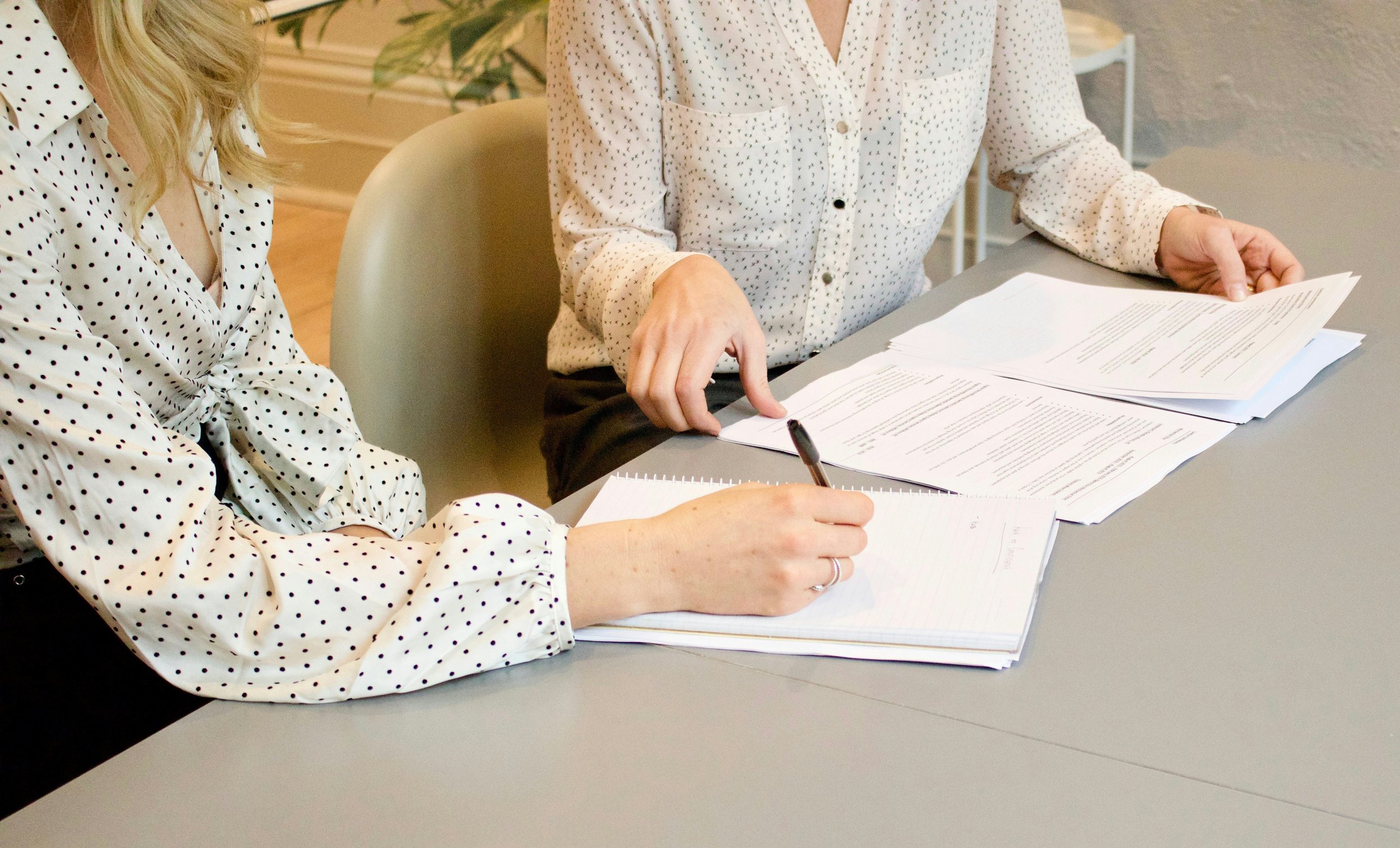 Two women seated at a table reviewing and discussing documents. One woman is taking notes, and the other is pointing at the documents. Both are wearing white shirts with black polka dots.