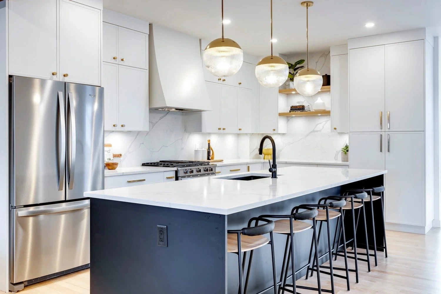 Modern kitchen with white cabinets, a large white marble island, a stainless steel refrigerator, black faucet, open wooden shelves, and three hanging pendant lights.