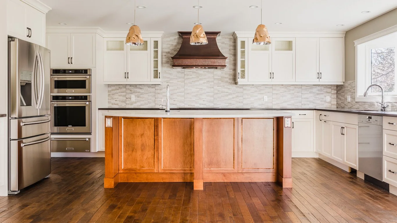 Modern kitchen with white cabinets, stainless steel refrigerator, oven, dishwasher, wooden floor, and a central island with a wood finish. There are hanging pendant lights above the island and a window next to the sink.