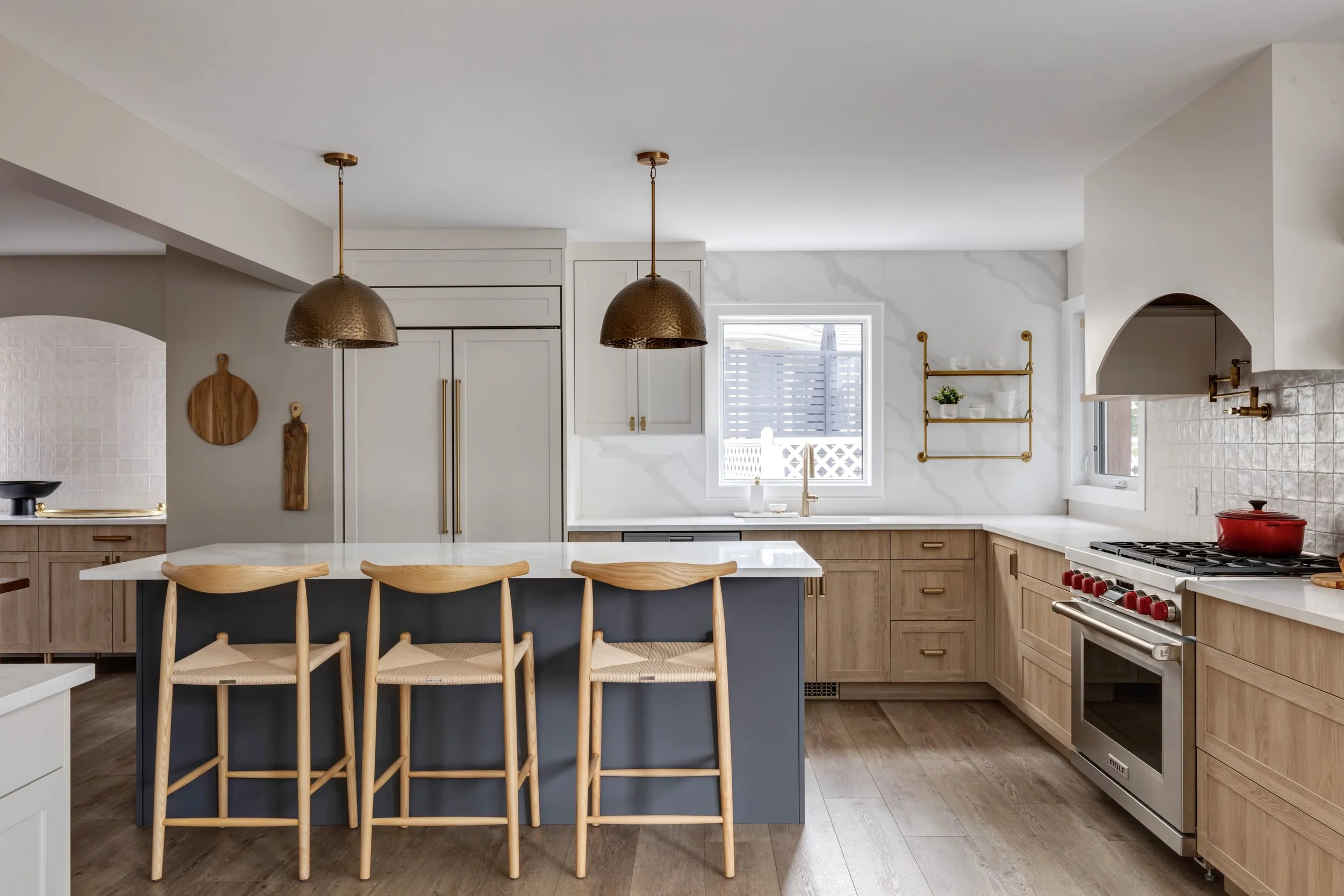 Modern kitchen with white cabinets, gold hardware, wooden accents, pendant lights over island, window with outdoor view, stainless steel stove, red kettle, wall-mounted shelving with plants and dishes.