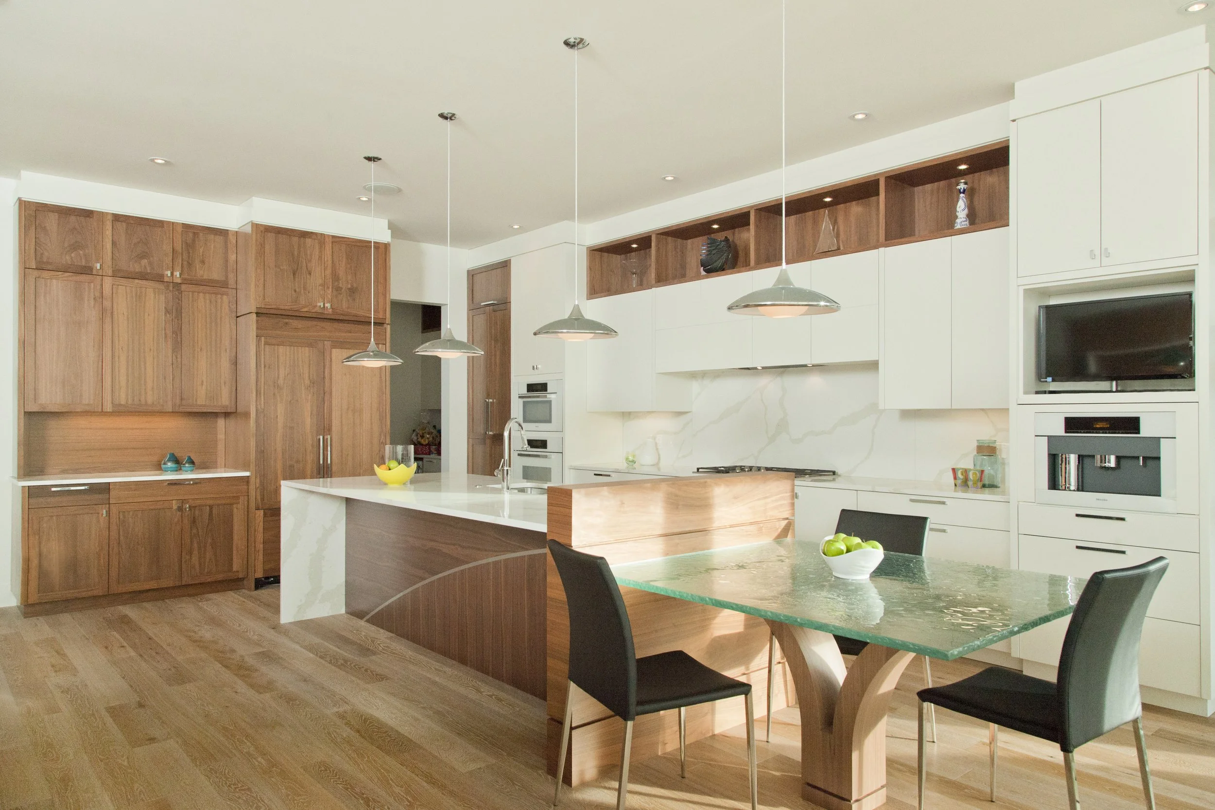 Modern kitchen with wooden and white cabinetry, a large kitchen island with a marble countertop, a glass-top dining table with green and black chairs, and decorative items on open shelves.