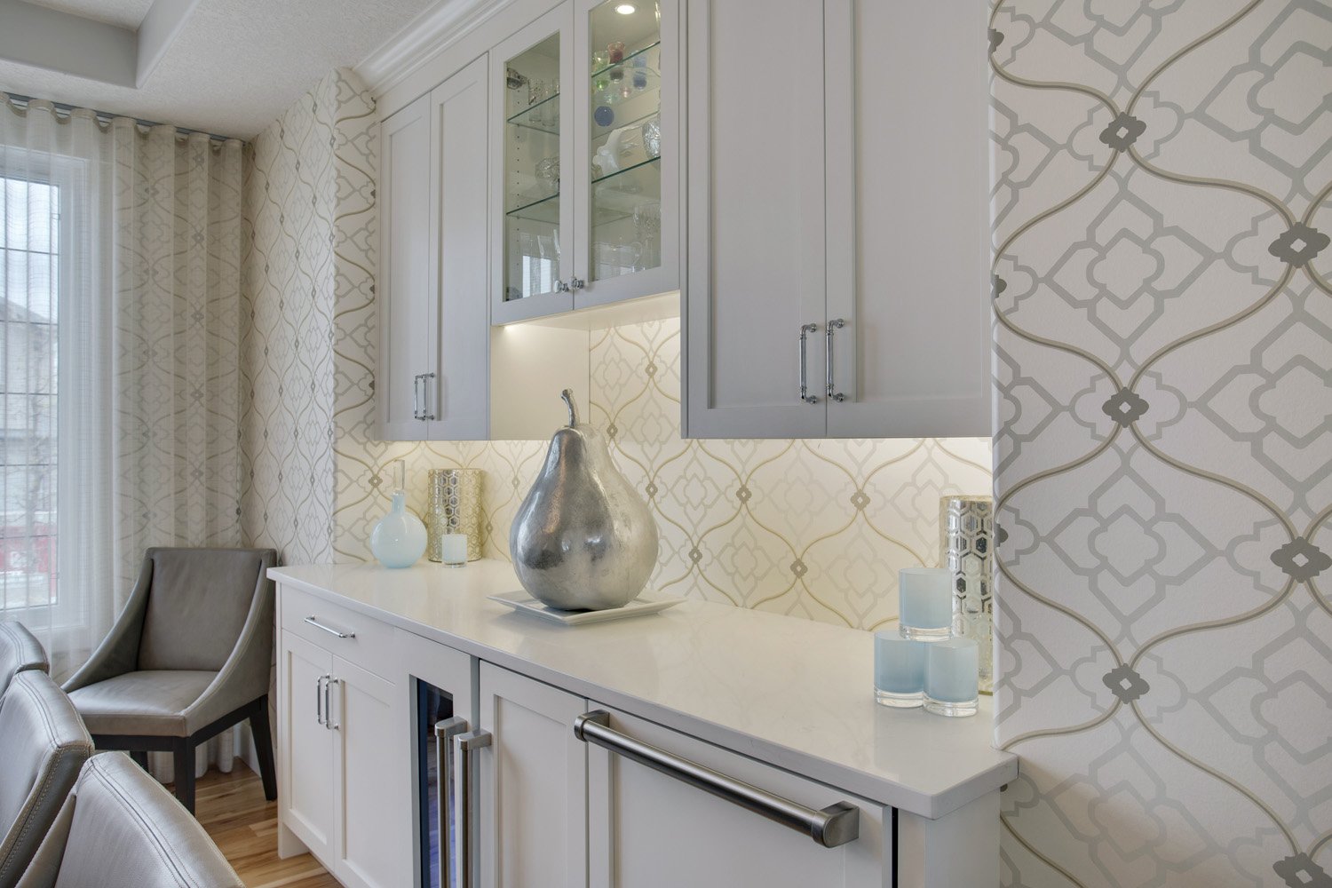 A modern kitchen sideboard with decorative vases and a large silver pear sculpture, surrounded by patterned wallpaper and glass-front cabinets.