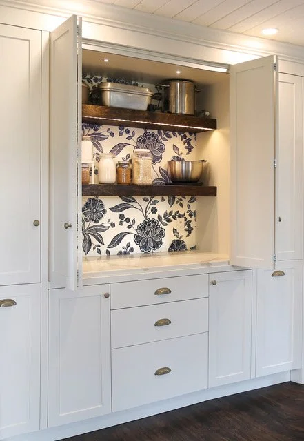 Open kitchen cabinet with white doors, showing two wooden shelves inside with various kitchen containers, bowls, and jars, and a black and white floral wallpaper background.