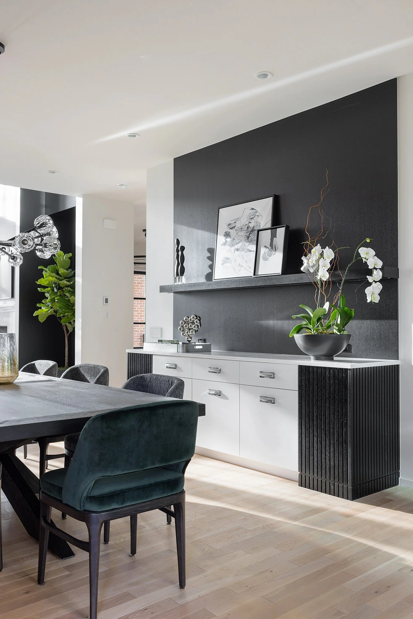 Modern dining room with black and white decor, including a black sideboard with framed artwork, a white orchid plant, and a dining table with dark green upholstered chairs.