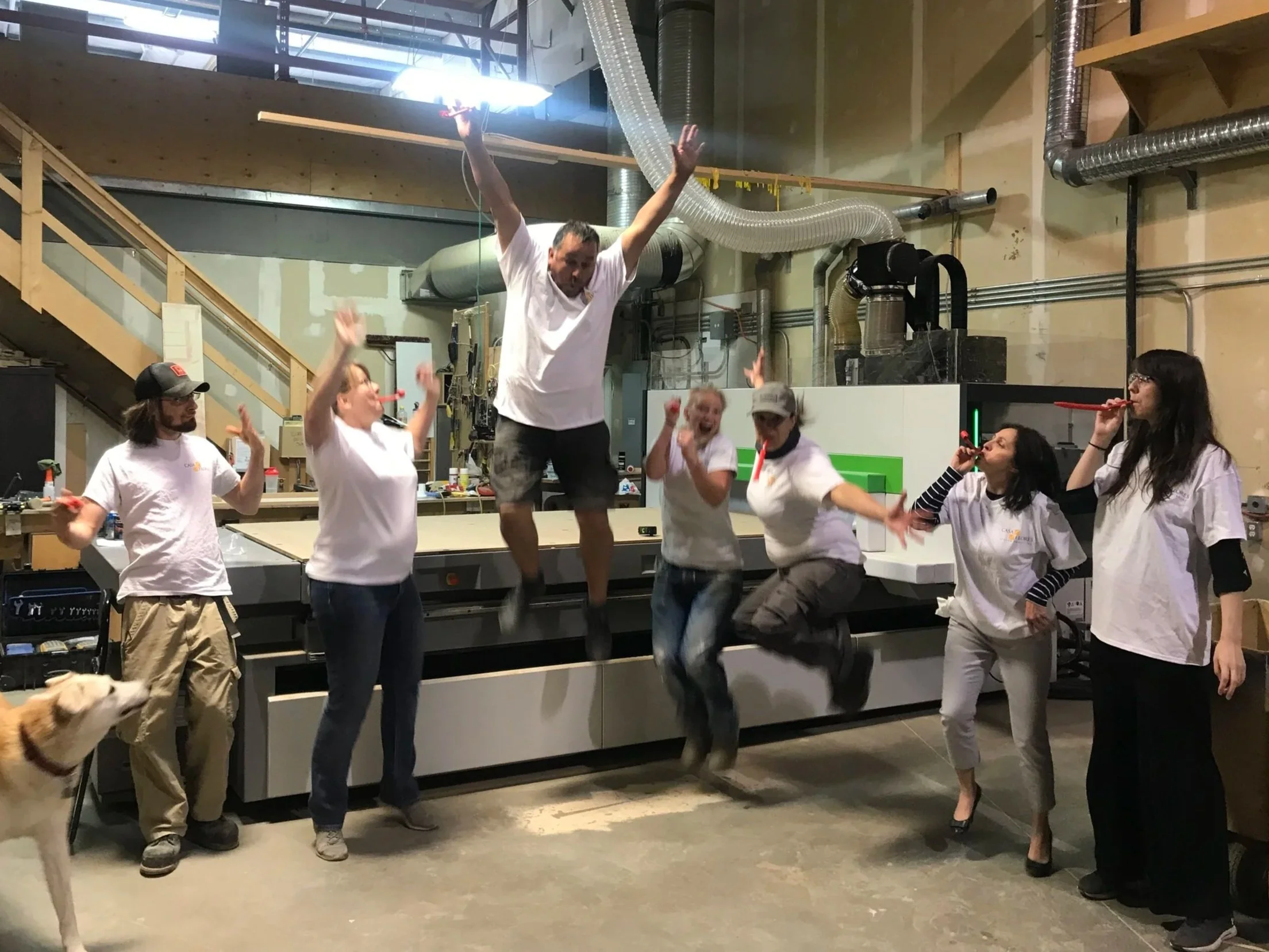 A group of people celebrating indoors, with some jumping and raising their arms. A woman in a black dress is standing nearby, holding a red party horn. There is a dog at the bottom left corner. The background includes industrial equipment and ventilation ducts.