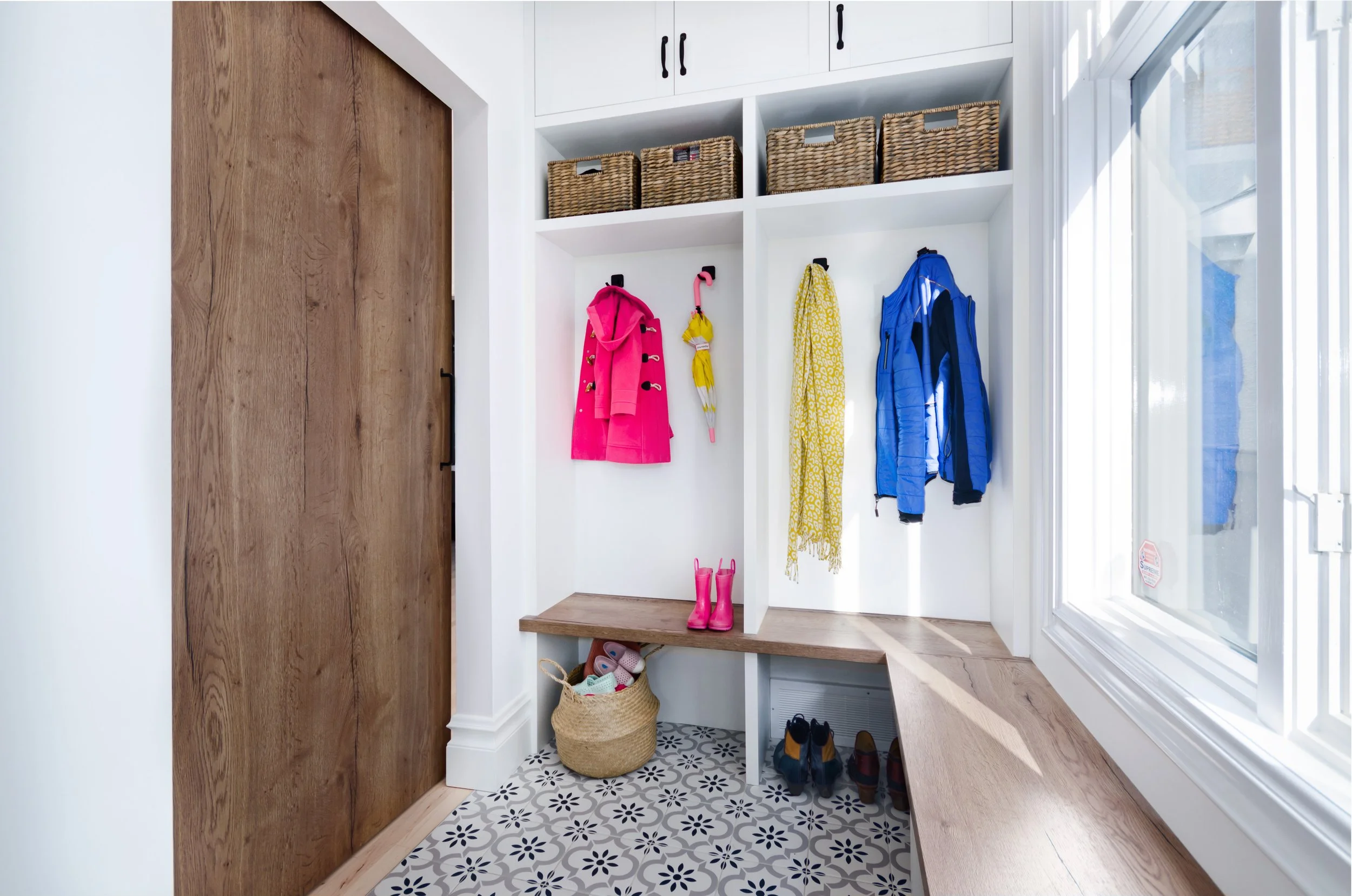 Entryway with white built-in cubbies holding baskets, colorful coats and scarves, pink rain boots on the wooden bench, shoes on the floor, and a window with natural sunlight.