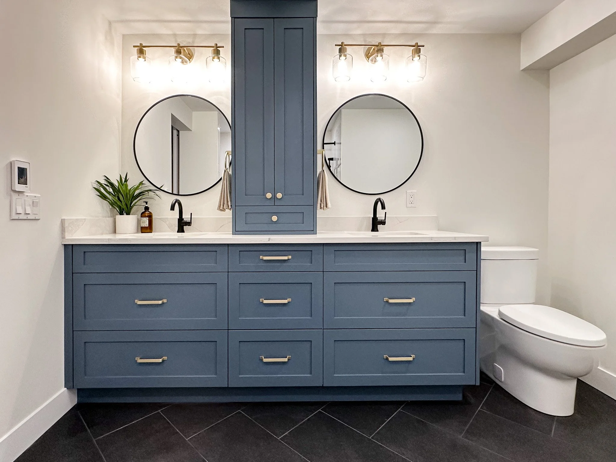 Modern bathroom vanity with blue cabinetry, two round mirrors, black faucets, a small green plant, and a beige towel. There is a white toilet beside the vanity.