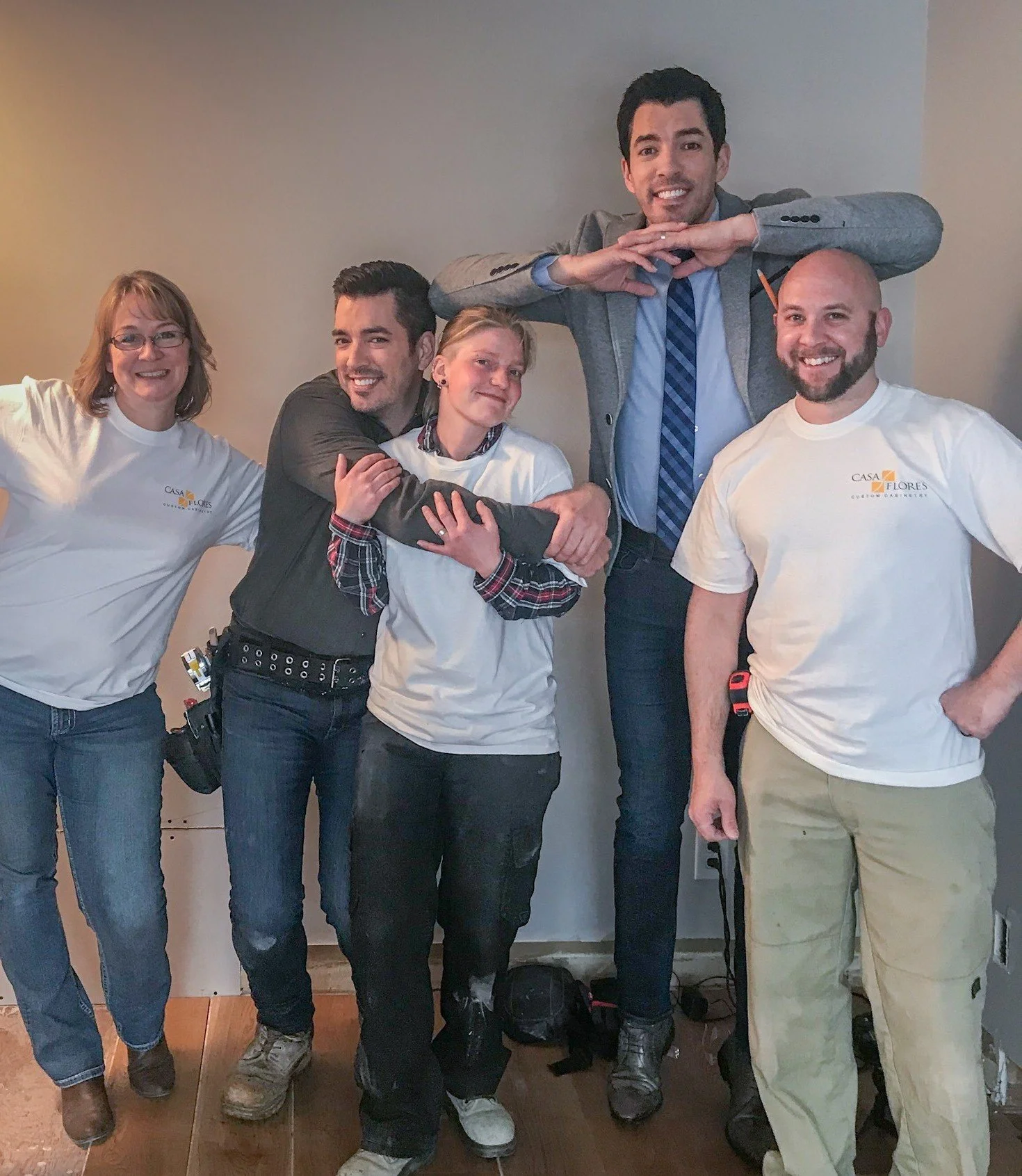 Group of five people standing inside a room, smiling, with some making playful gestures; two women and three men, dressed casually and professionally, with one man in a business suit.
