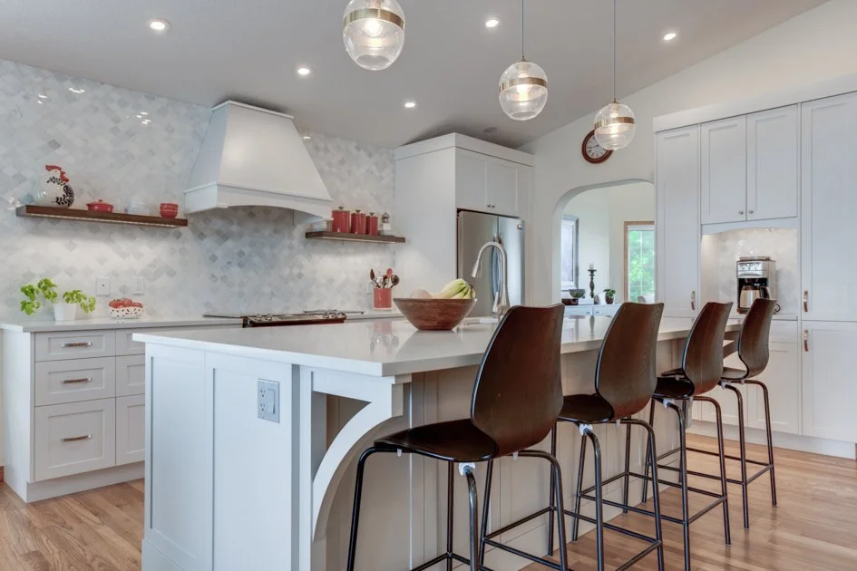 Modern kitchen with white cabinets, a large island with barstools, stainless steel appliances, decorative red accents, and pendant lights.