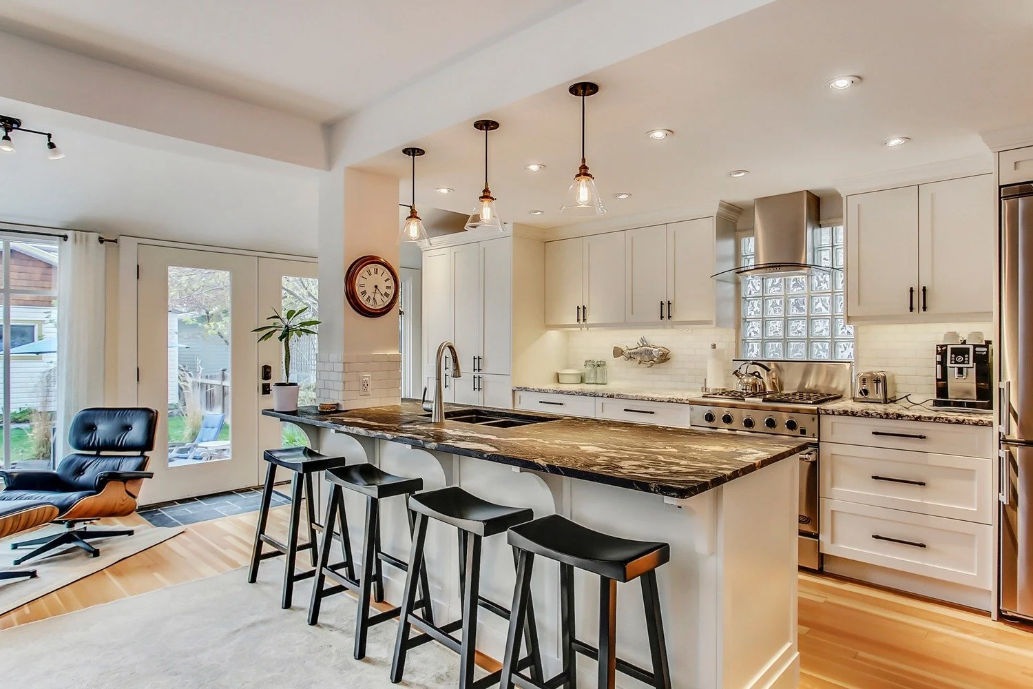 Modern kitchen with white cabinets, stainless steel appliances, granite countertops, and a breakfast bar with four black stools. There is a wall clock, pendant lights, and a plant on the counter. Sliding glass doors lead to an outdoor patio.