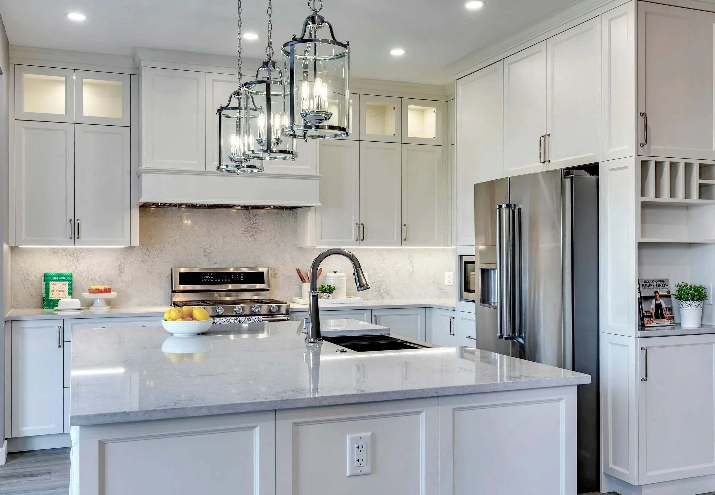 A modern white kitchen with a large stone island in the center. The kitchen has white cabinets, a stainless steel refrigerator, and a marble backsplash. There are hanging pendant lights above the island, and various small items, including a bowl of lemons, books, and decorative plants, are placed on the countertop.