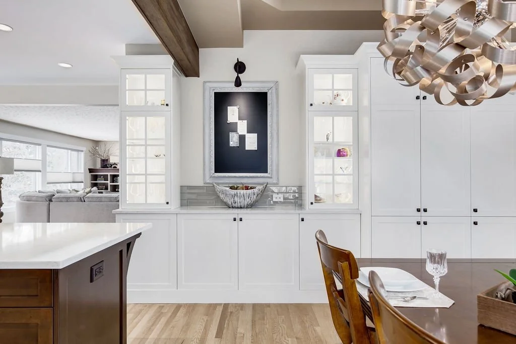 Modern kitchen with white cabinetry, a dark wooden beam on the ceiling, a black chalkboard on the wall, and a dining table set with glasses and utensils.