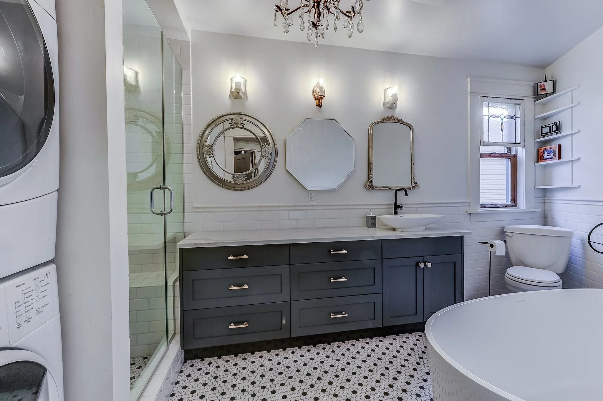 A modern bathroom with a white and gray color scheme, featuring a black vanity with gold handles, a round bathtub, a toilet, and a shower with glass doors. There are three decorative mirrors and artwork on the wall, a window with white trim, and a ch