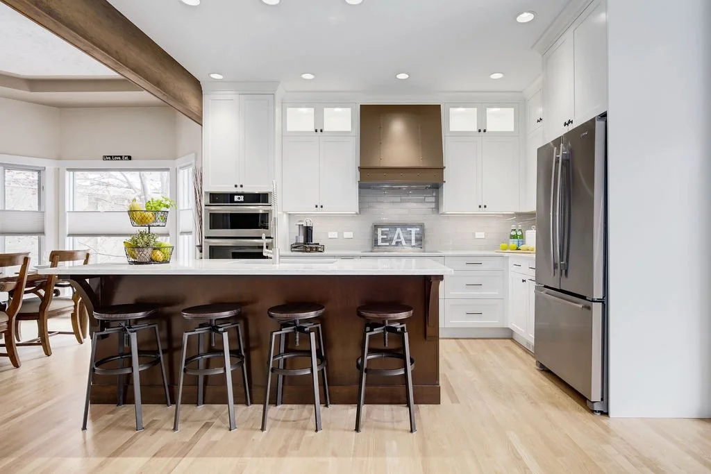 Modern kitchen with white cabinets, stainless steel refrigerator, built-in oven, gray backsplash, and a large brown island with four black stools. There is a decorative sign that says "EAT" on the countertop.