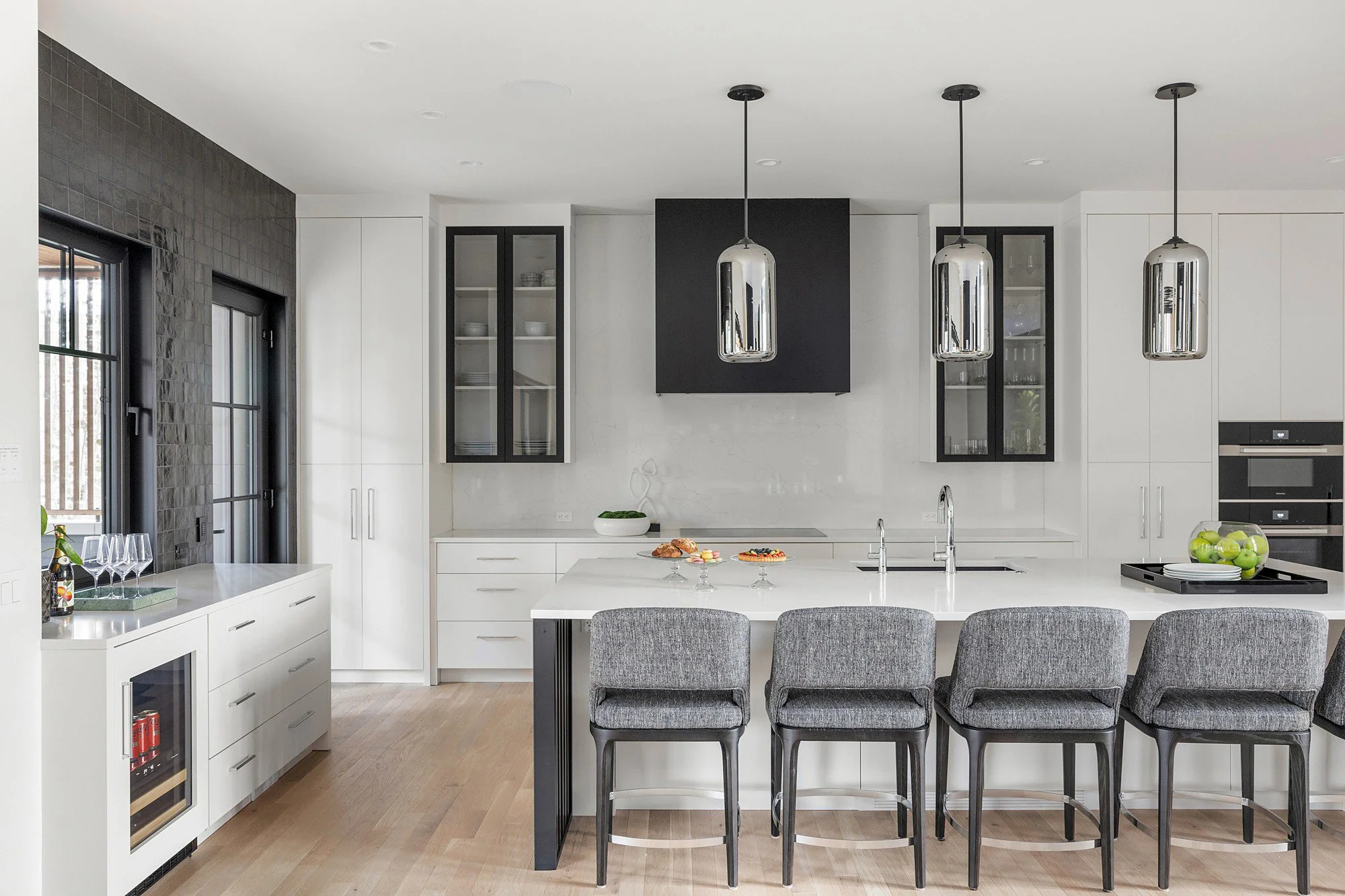 Modern kitchen with black and white cabinetry, stainless steel pendant lights, and a breakfast bar with gray upholstered chairs. There are display cabinets with glass doors, a matte black square feature wall, and a window with black frames.