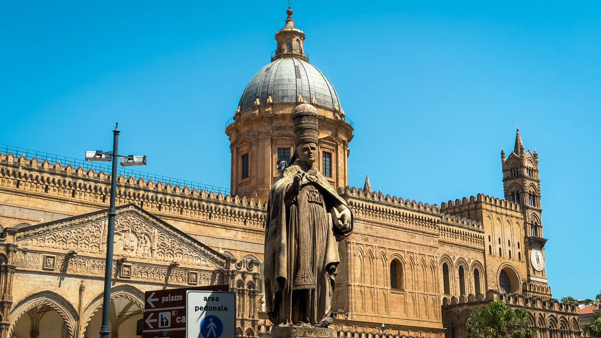 Palermo Cathedral with its dome, crenellated walls and a stone statue in the foreground under a blue sky.