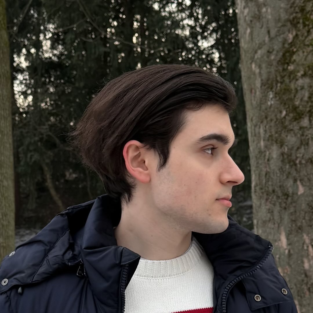 Profile of a young man with dark hair, wearing a black jacket and white sweater, standing outdoors near trees.