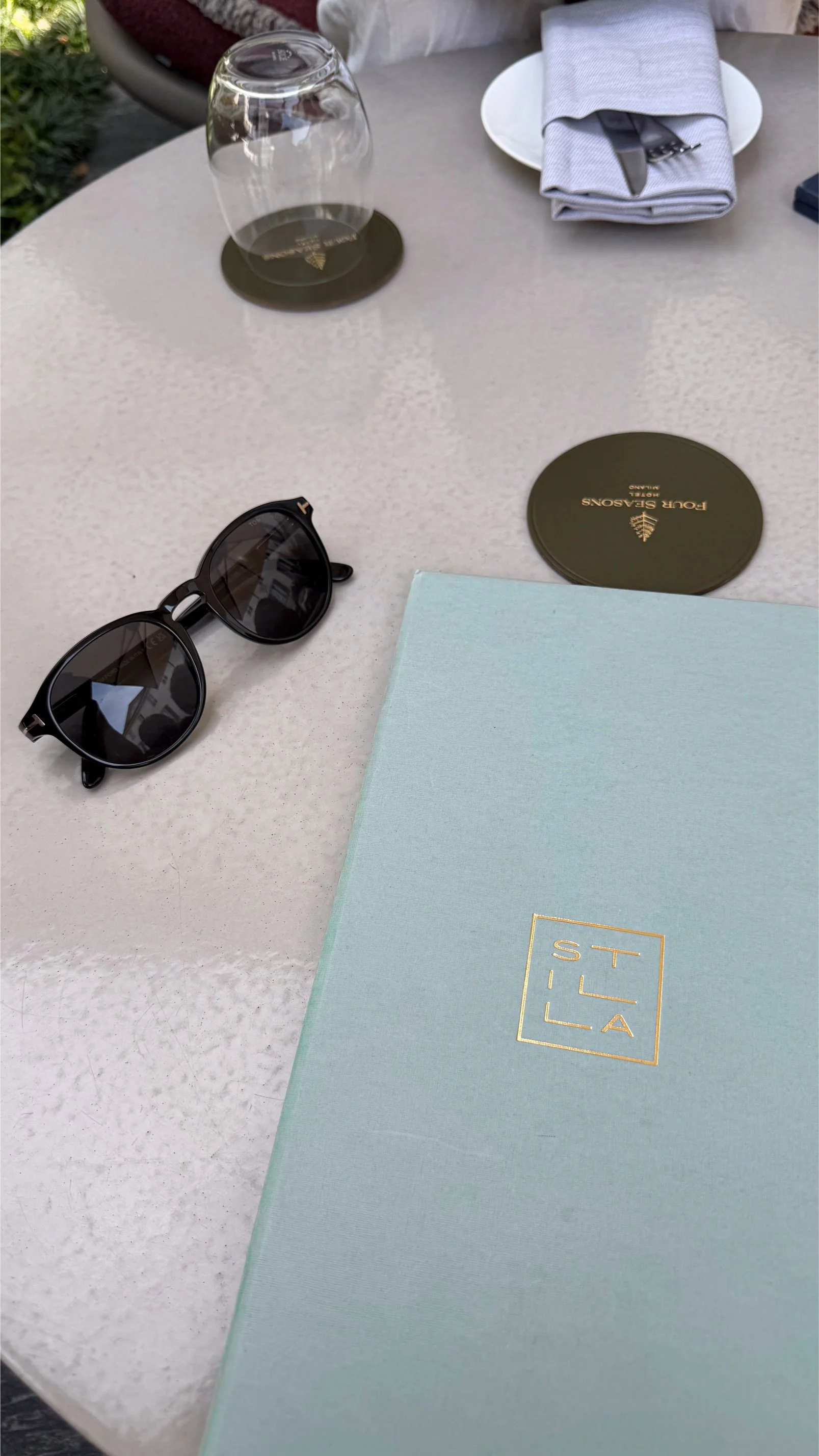 A table with a pair of sunglasses, a blue notebook, a set of cutlery wrapped in a napkin, a glass upside down on a coaster, and a round container with gold text. The table is outside.