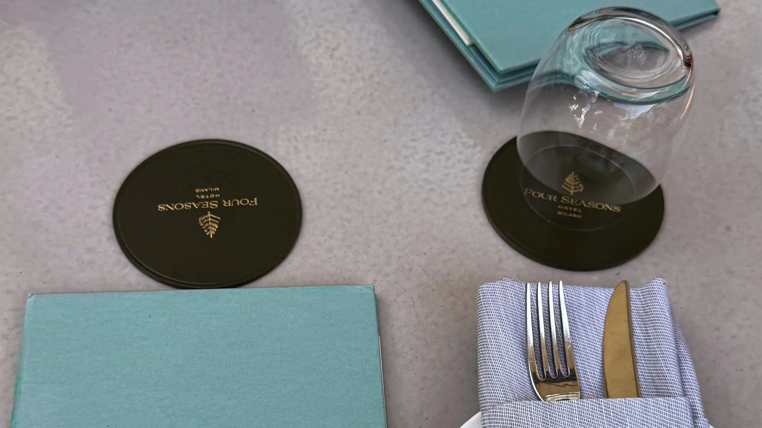 Breakfast table setting at Stilla Garden, Four Seasons Hotel Milano, with branded coasters, glassware, cutlery and turquoise menus.