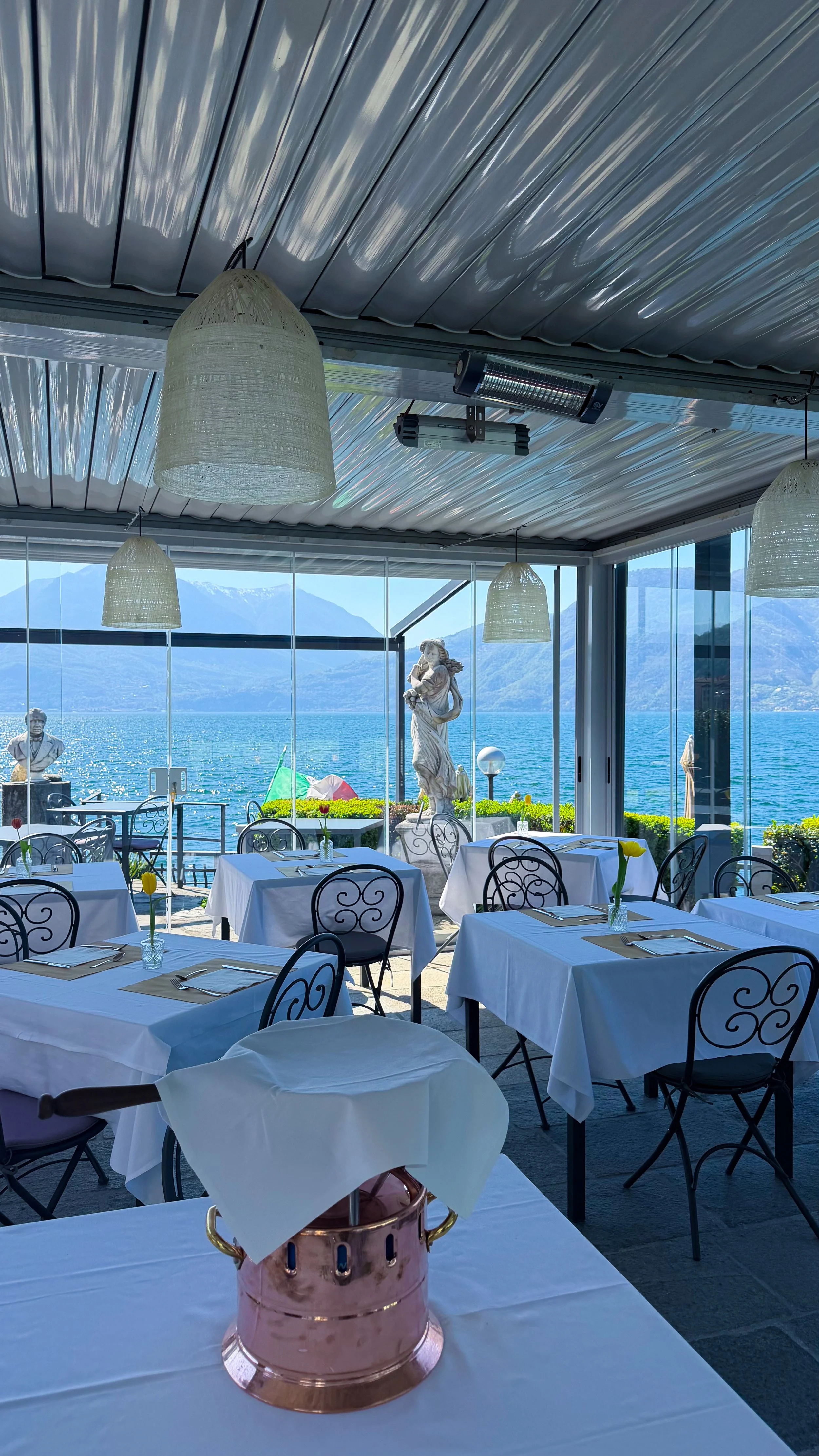 Restaurant interior overlooking Lake Como, with white table settings, glass windows, classical statues, and mountain views across the water.