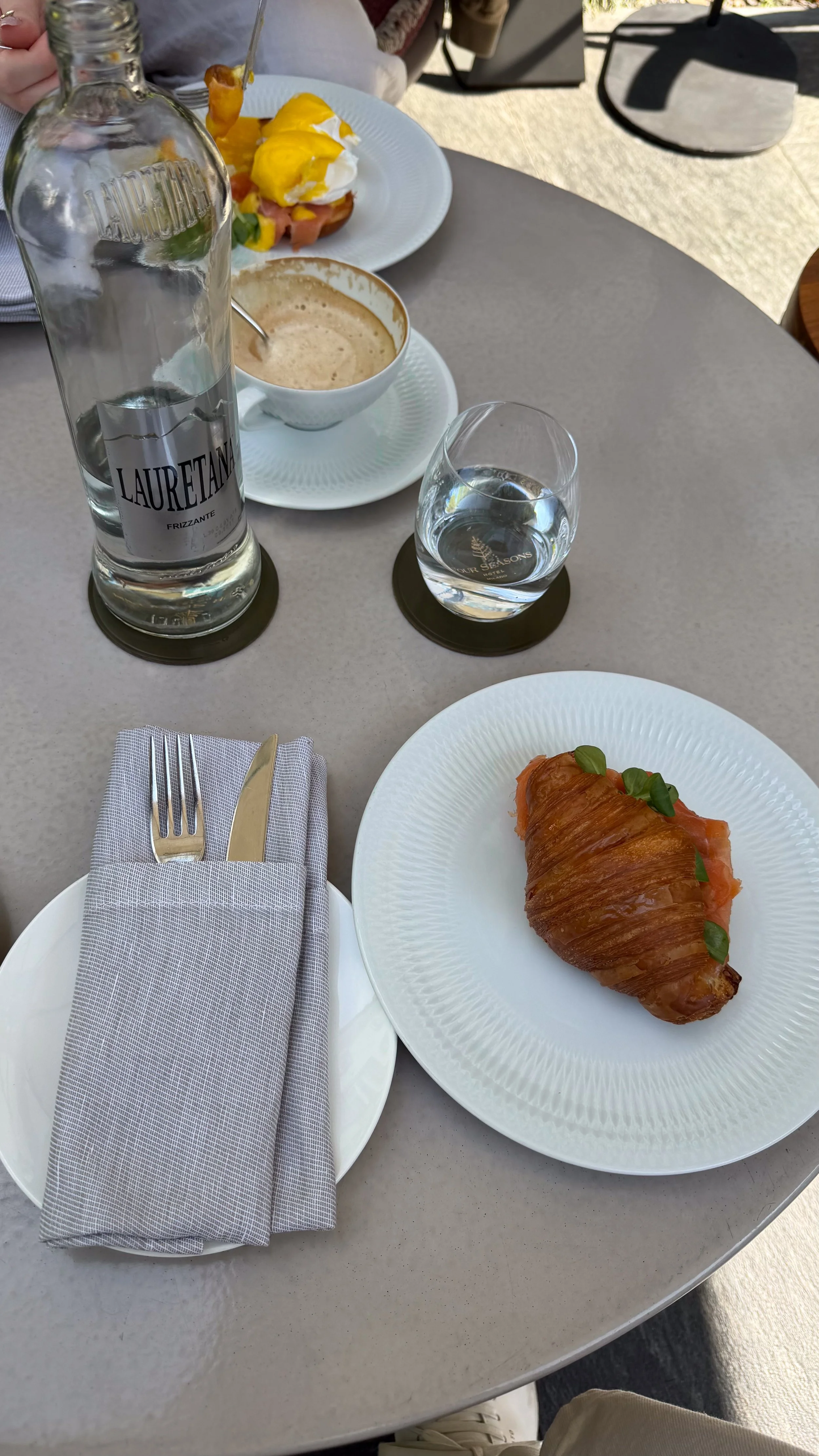 A table with a croissant on a white plate, a glass of water, a bottle of water, and a cup of coffee, with eggs Benedict in the background.