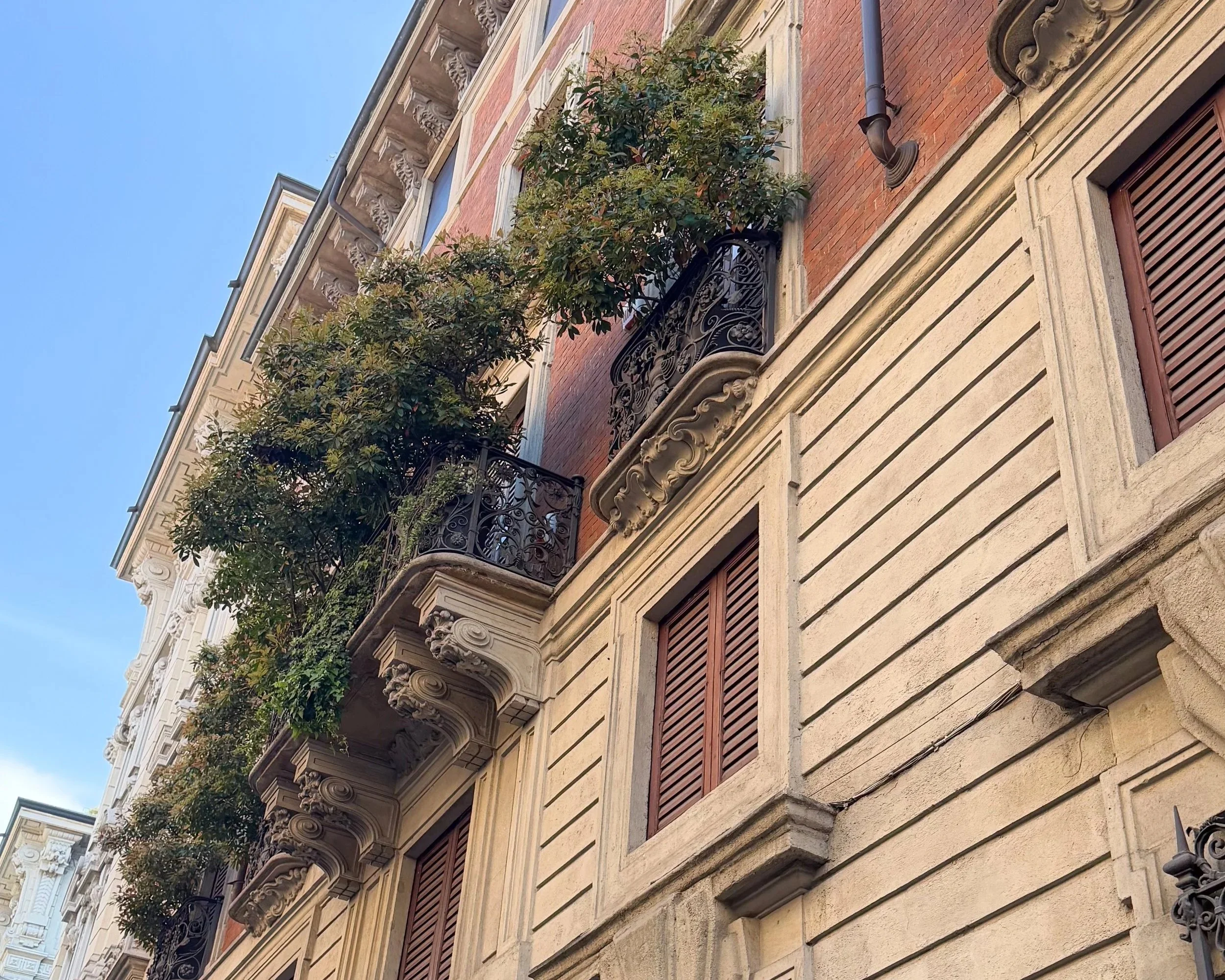 Historic Milan building facade with ornate balconies, shutters, and greenery against a blue sky.